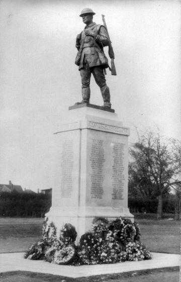 Black and white photograph. A community cenotaph. A large cement pillar, with lists of names engraved on each side. On top, a life-size soldier in mid step, rifle slung on his back, looks strongly into the distance. Wreaths are piled at the base.